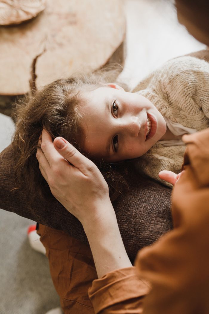 gallery-4 A child lies on a pillow, comforted by a parent's touch. Warm and intimate indoor setting.