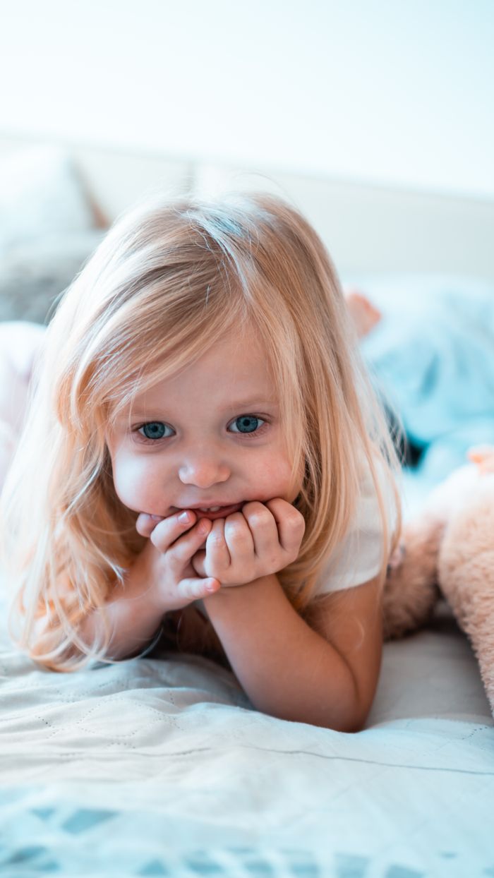 gallery-1 Adorable blonde-haired child with blue eyes lying on a bed, looking content and peaceful.